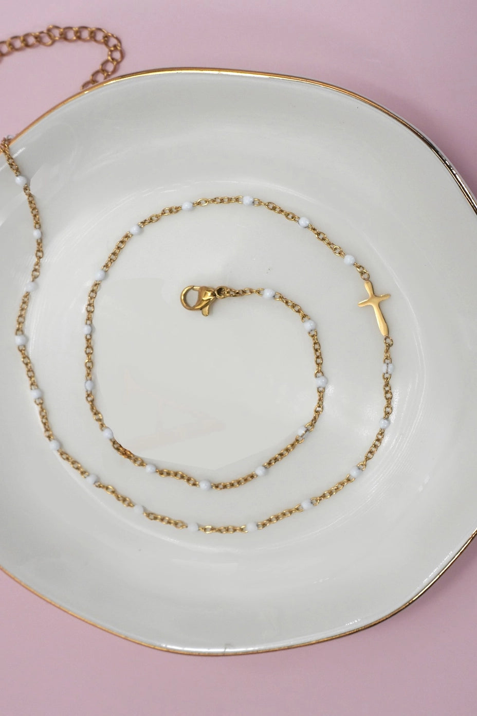 White beaded gold cross charm stainless steel necklace on ceramic plate with pink background