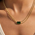 A close-up of a model's neck and chest wearing a double-strand gold chain necklace with a small rectangular green emerald-cut center stone.
