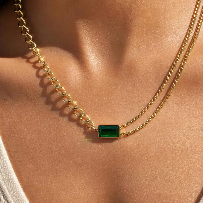 A close-up of a model's neck and chest wearing a double-strand gold chain necklace with a small rectangular green emerald-cut center stone.