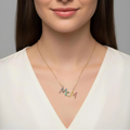 A woman with long dark brown hair is shown from the chest up against a plain gray studio background, wearing a delicate gold necklace with the name 'MCM' in colorful lettering.