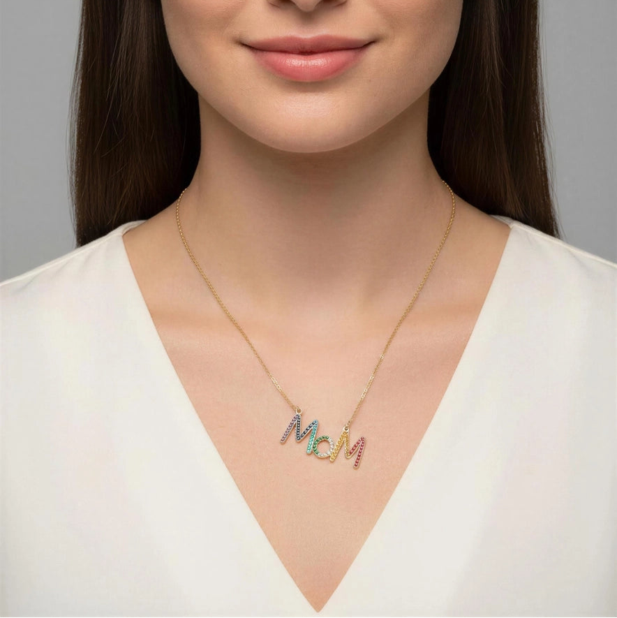 A woman with long dark brown hair is shown from the chest up against a plain gray studio background, wearing a delicate gold necklace with the name 'MCM' in colorful lettering.